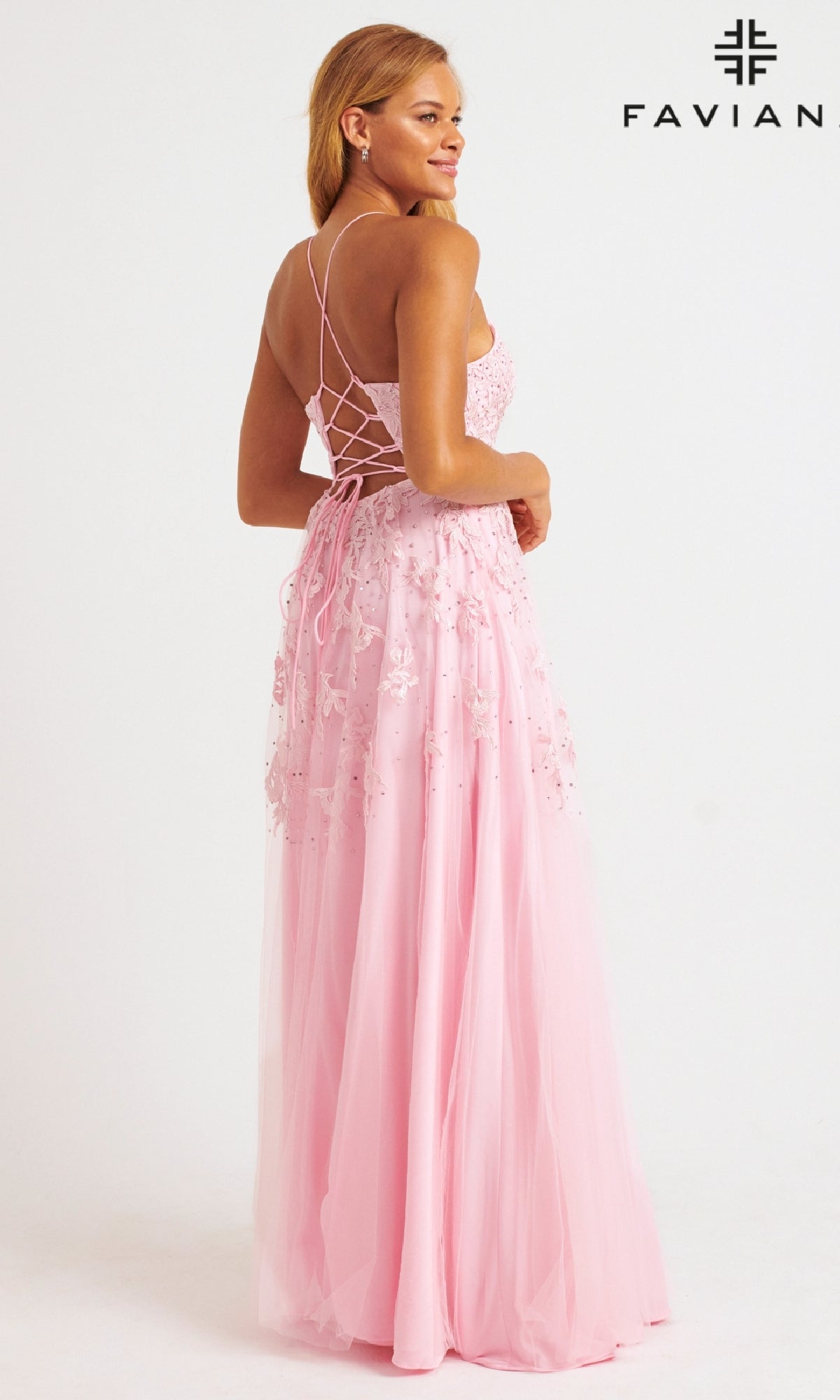 A woman stands looking over her shoulder in a long, light pink Faviana Embroidered A-Line Long Prom Dress with a lace-up back and 3D flower embroidery.